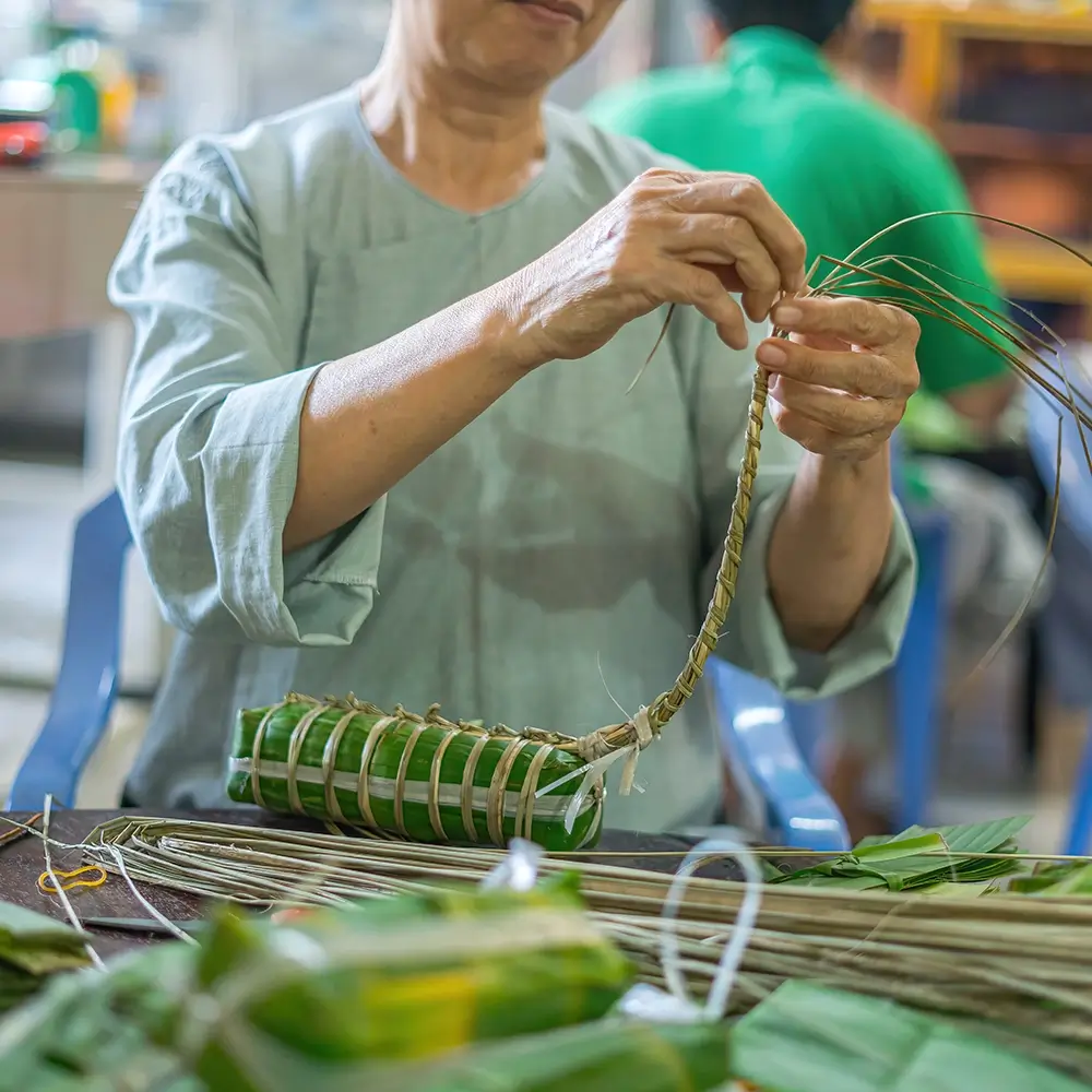 focus-old-woman-with-traditional-vietnamese-dress-ao-ba-ba-making-wrapping-tet-cake-vietnamese-lunar-new-year-tet-food-outdoor-by-hands copy